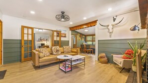 A spacious living room with a sofa, coffee table, and a dining area. - Cozy Cabin w/ Hot Tub, Game Room & Private Writer's Nook - Close to town! (Evergreen)
