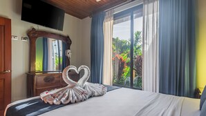 A hotel room with a large bed, a heart-shaped towel arrangement, a wooden dresser, a flat-screen TV, and a window with curtains. - Kaluma Frog Farmstay Villa La Fortuna (San Ramon)