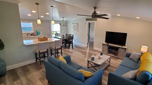 A modern living room with a sofa, coffee table, and TV. There is a dining area with chairs and a table, and a kitchen with a window and counter. - St Augustine waterfront home on canal w/ private dock, walk to beach! (St. Augustine)