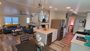 A modern living room with a sofa, coffee table, and dining area. There is a kitchen with a refrigerator, stove, and sink. A ceiling fan and pendant lights are visible. - St Augustine waterfront home on canal w/ private dock, walk to beach! (St. Augustine)