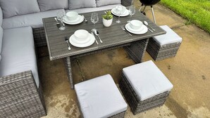 An outdoor dining area with a gray wicker sofa, a dark wooden table set with white plates, bowls, and cutlery, and a black grill in the background. - Fast Wi-Fi/Comfy beds/Free parking/3bdr house/4TVs
 (Aurora)