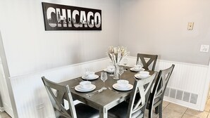 A dining room with a table set for six, white plates, glasses, and a vase with dried flowers. - Fast Wi-Fi/Comfy beds/Free parking/3bdr house/4TVs
 (Aurora)