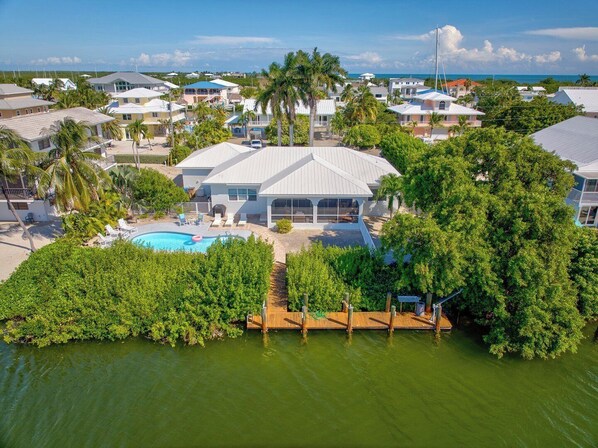 A residential area with a large house featuring a swimming pool, surrounded by lush greenery and a view of the ocean.