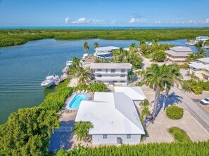 An aerial view of a coastal property with a swimming pool, multiple buildings, and boats docked near the water.