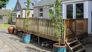 Een woonhuis met een houten terras, twee potplanten, en een gebouw met een grijs dak en een witte gevel.