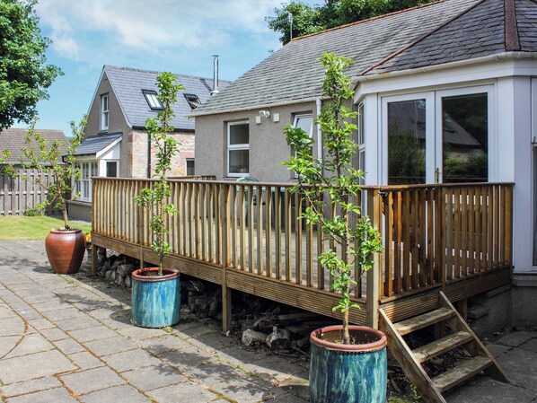 A residential property with a wooden deck, two potted plants, and a house with a grey roof and white exterior. - Pine Tree Cottage - UK45925 (Ballater)