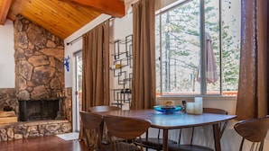 A cozy room with a stone fireplace, wooden ceiling beams, a dining table with chairs, and a large window with curtains. - Cozy Mountain Getaway in Arrowbear Lake, minutes from ski resorts. Pet-Friendly! (Running Springs)