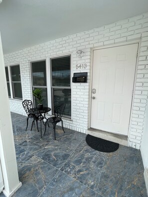 Une façade en briques blanches avec une boîte aux lettres, une porte et un petit espace patio comprenant une table et des chaises.