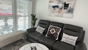Una sala de estar con un sofá de cuero negro, dos almohadas con el texto 'welcome home', una mesa de centro de mármol y un cuadro abstracto grande en la pared.