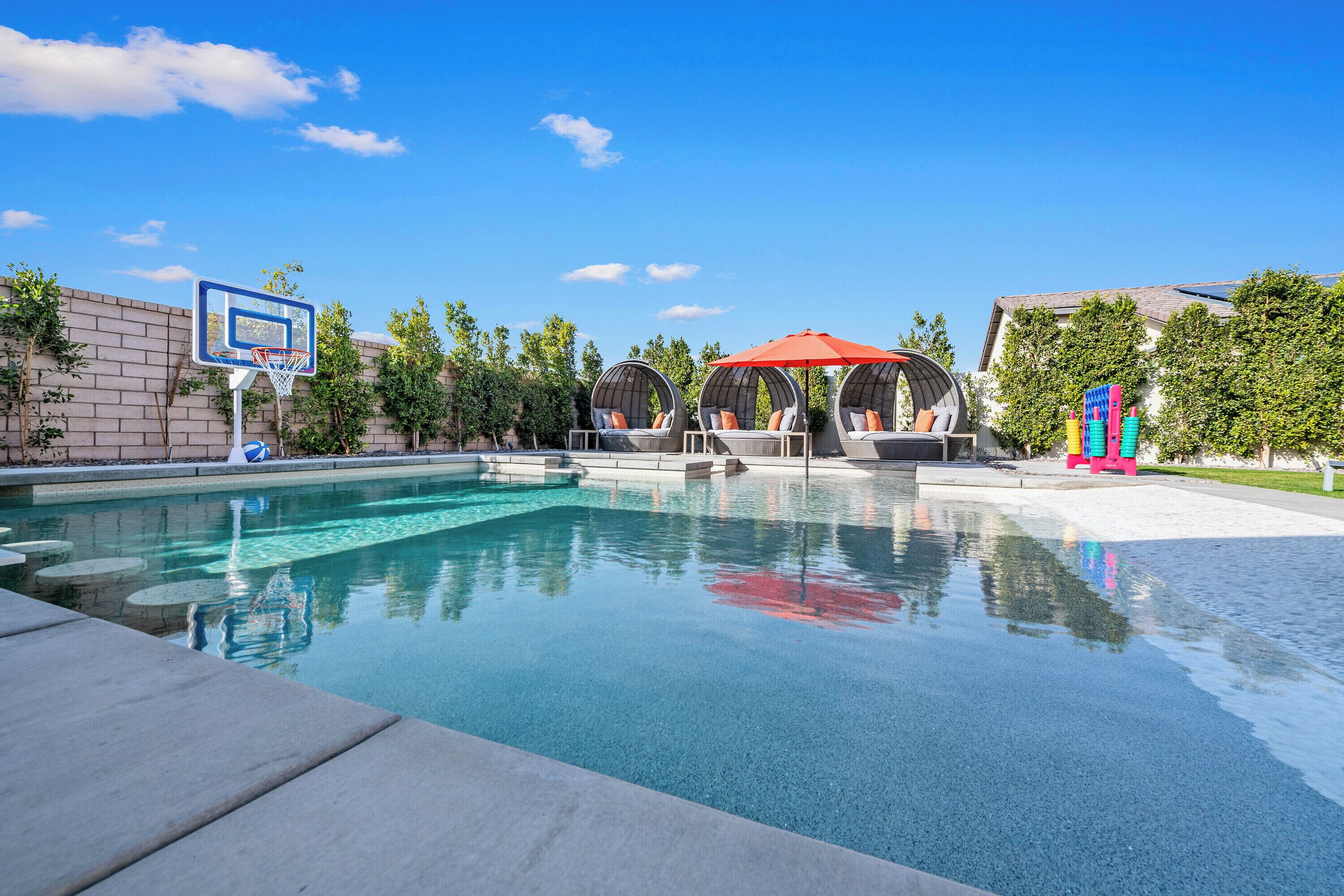 A swimming pool with a basketball hoop, lounge chairs, and umbrellas.