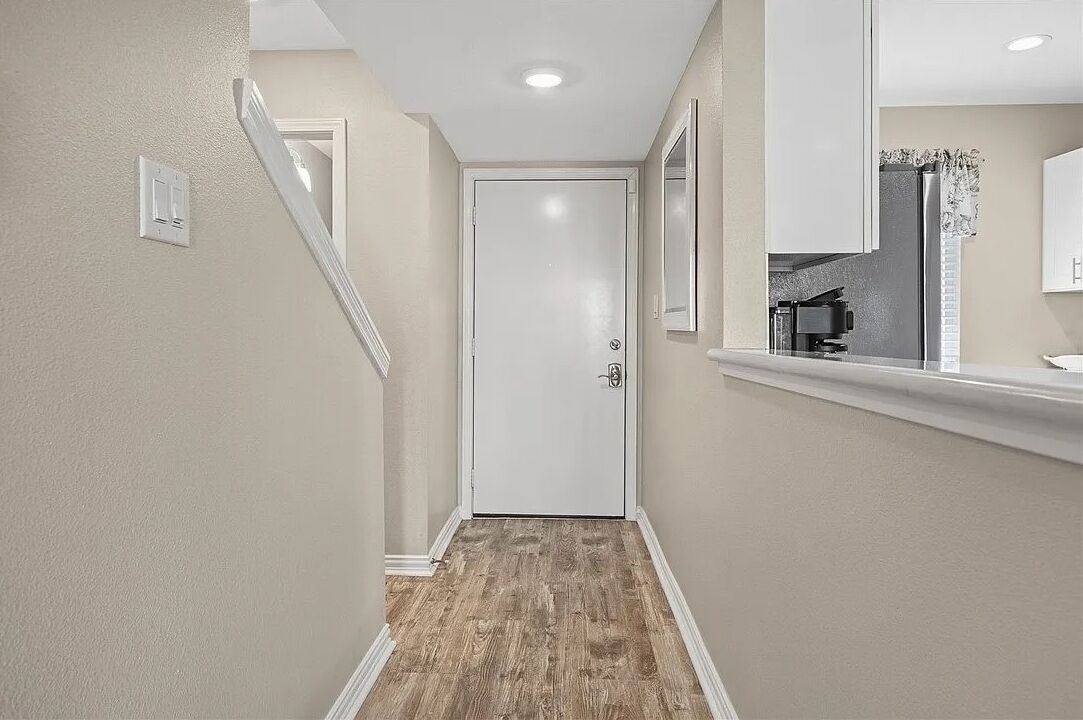 Um corredor estreito com piso de madeira, uma porta branca e uma área de cozinha com geladeira e pia.