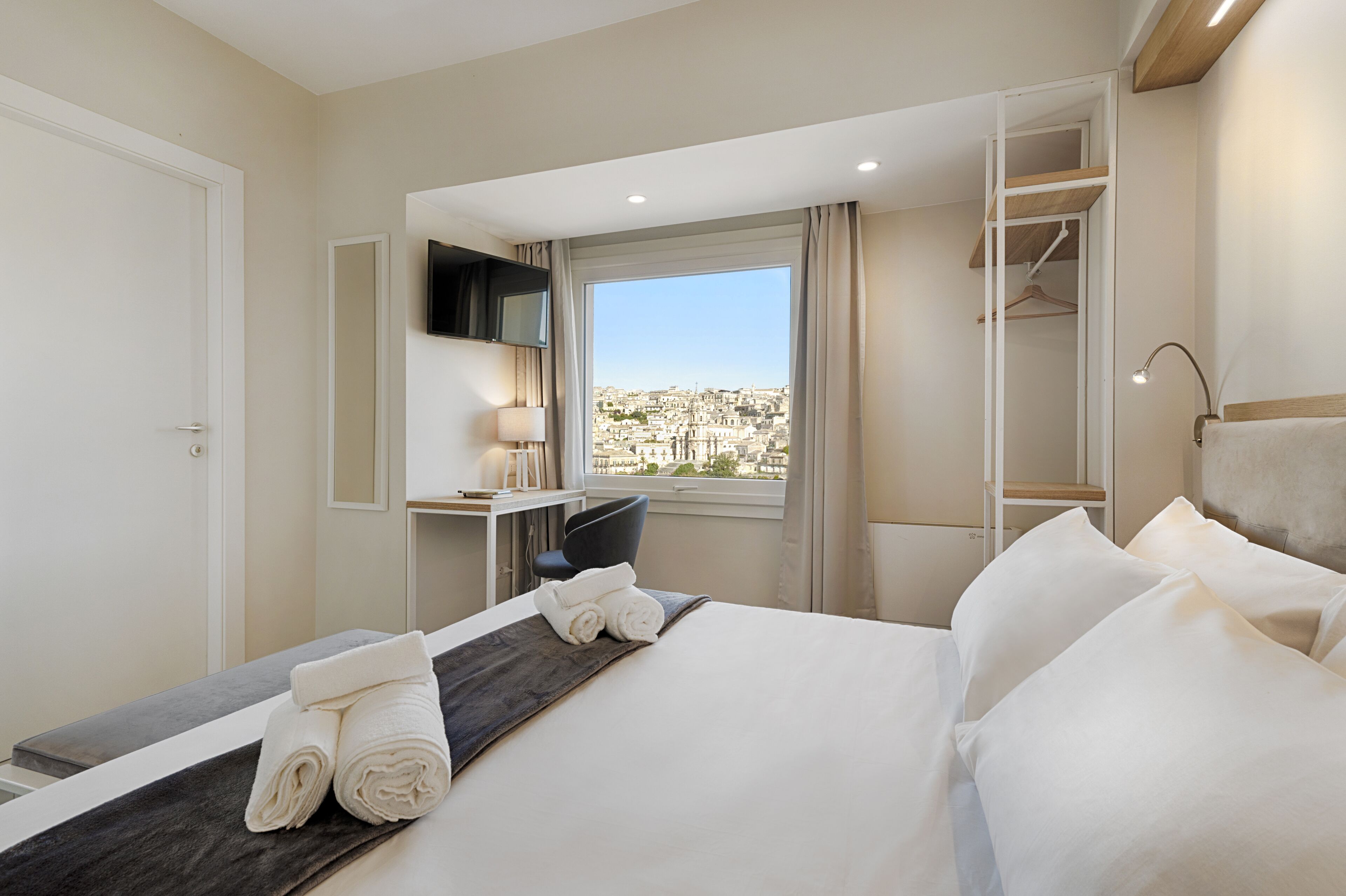 A hotel room with a bed, a desk, a chair, a TV, and a window with a city view.
