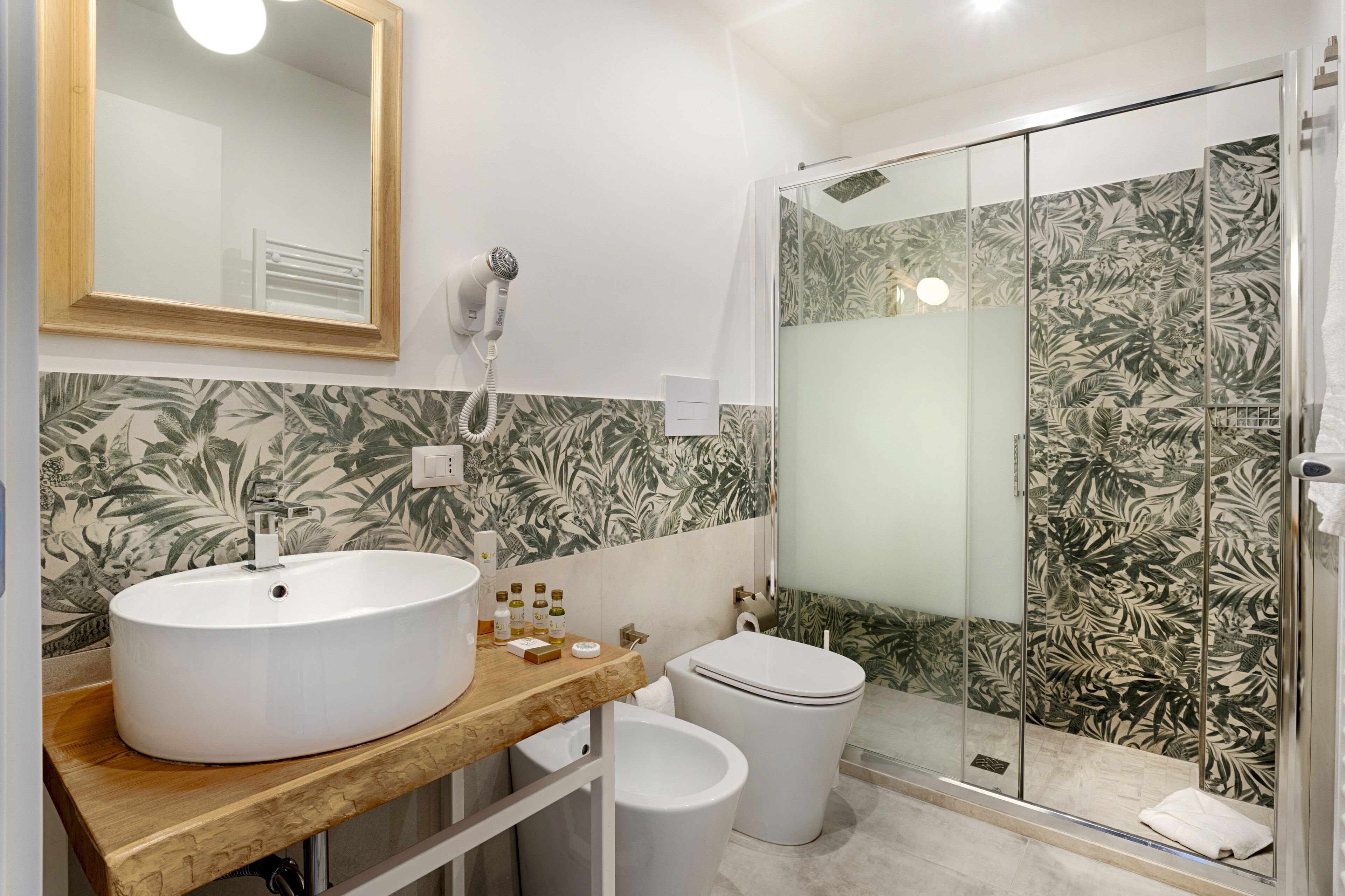 Une salle de bain avec un meuble-lavabo en bois, un lavabo blanc, des toilettes et une douche en verre avec un pare-douche en carreaux décorés de motifs de feuilles.