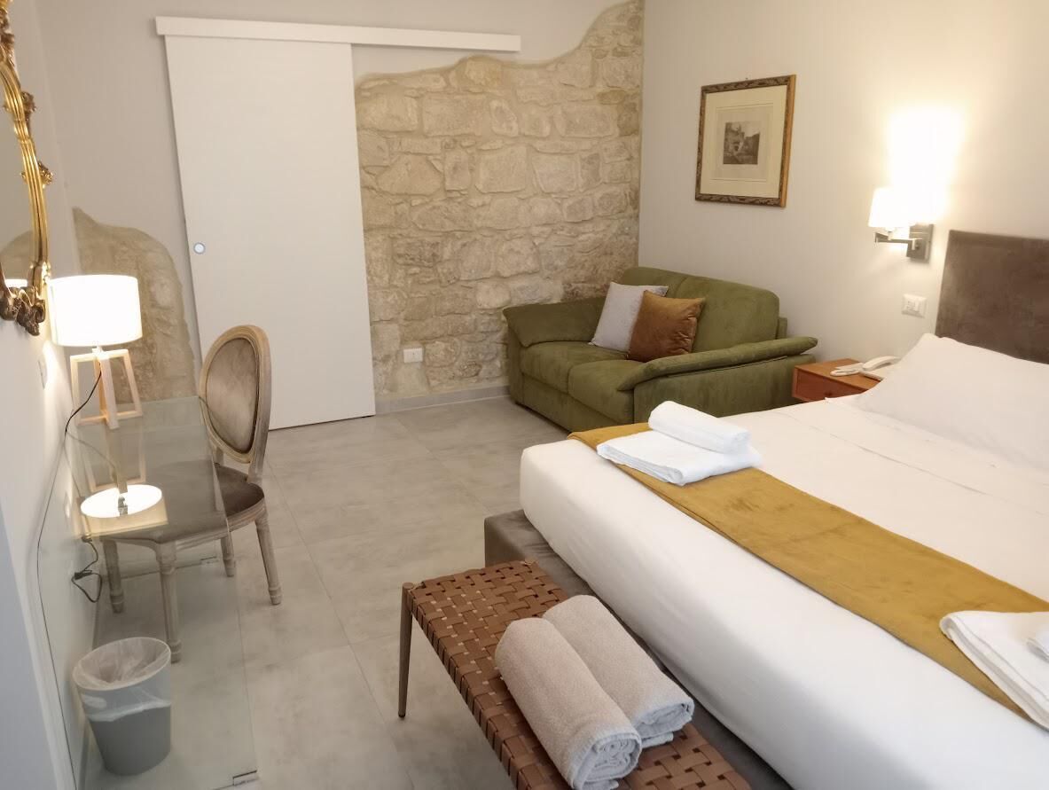 Habitación de hotel con cama, sofá verde, banco de madera, una pequeña mesa con silla y un cubo de basura.
