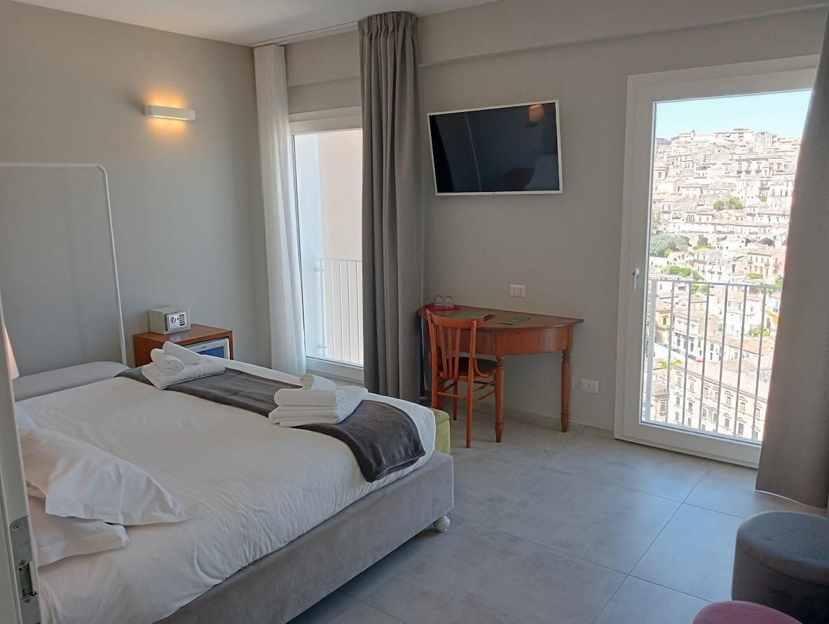 Habitación de hotel con cama, escritorio, televisor y balcón con vistas a la ciudad.