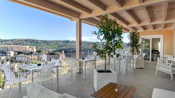 A covered outdoor terrace with white furniture, a wooden table, and a potted plant.
