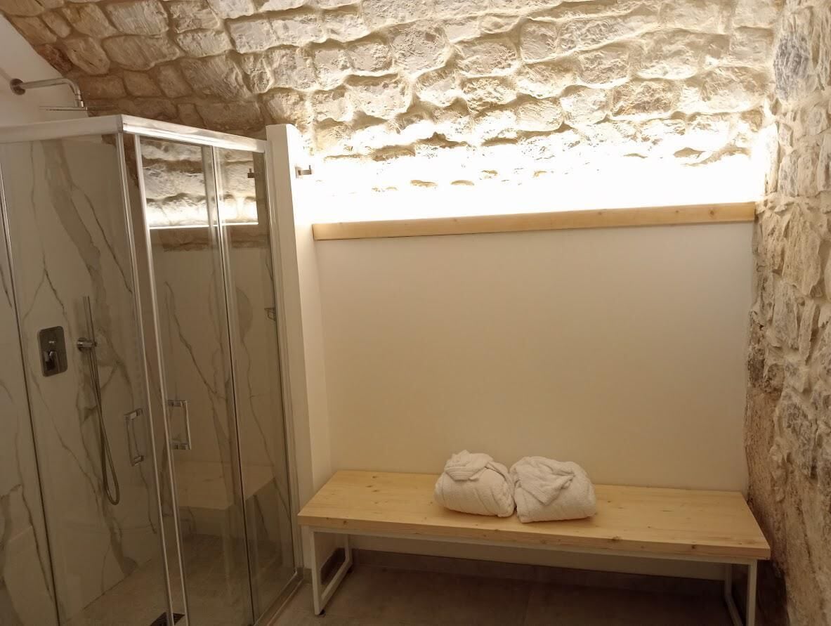 A bathroom with a glass-enclosed shower, a bench with towels, and a stone wall.