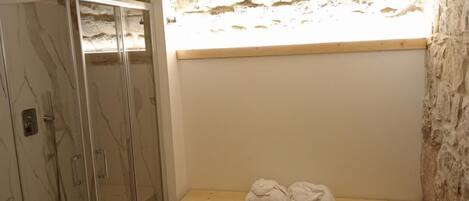 A bathroom with a glass-enclosed shower, a bench with towels, and a stone wall.