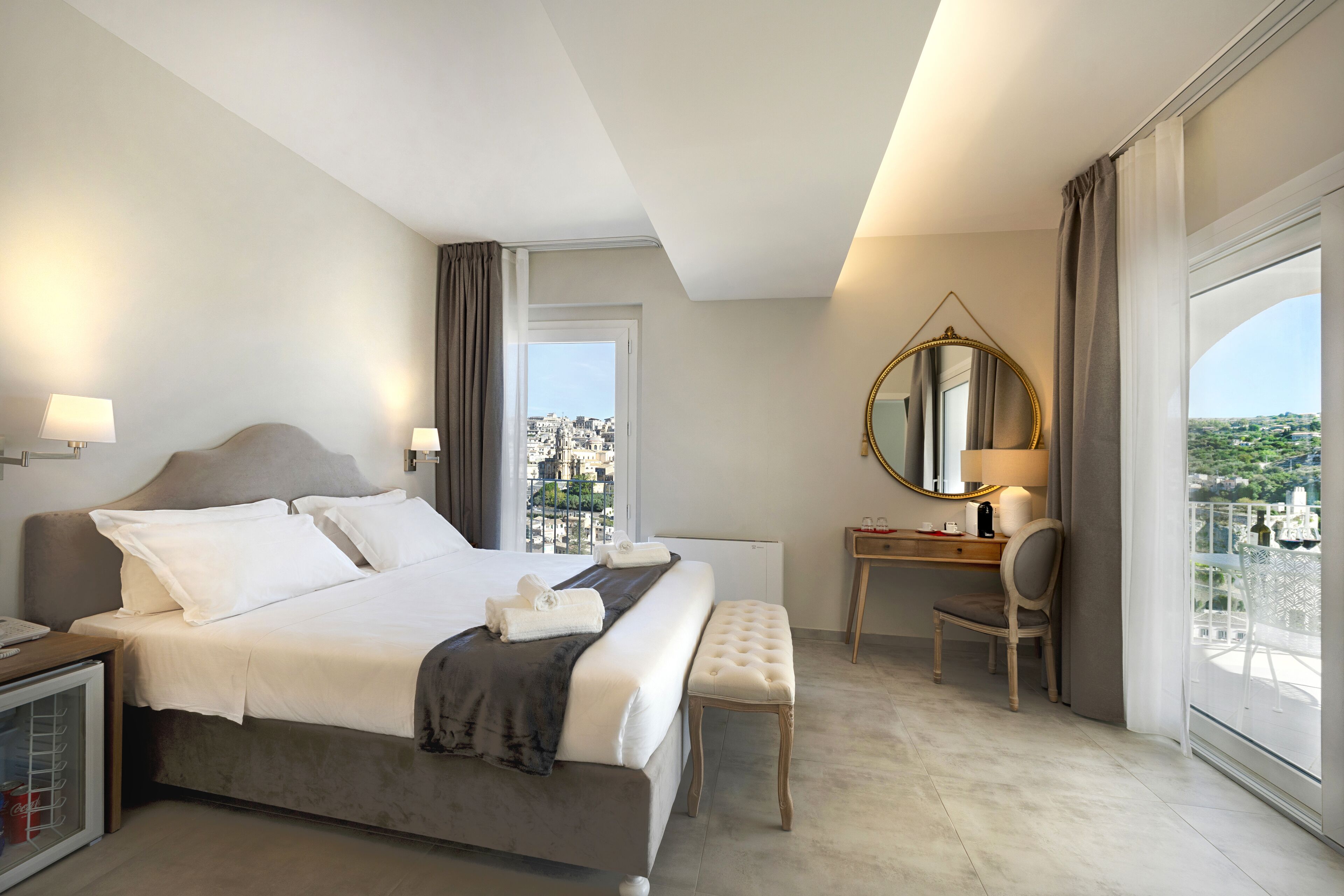 A hotel room with a large bed, a desk, a chair, a mirror, and a view of the city.