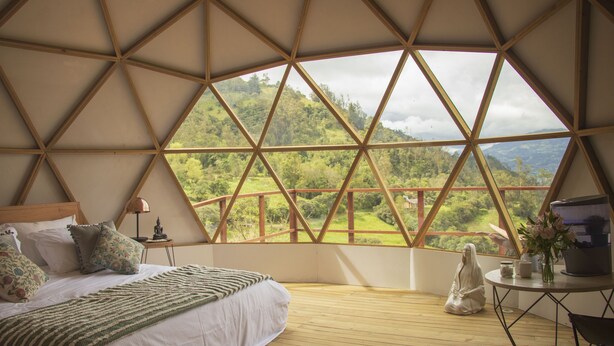 A spacious bedroom with a large bed, a nightstand, and a view of a lush landscape through a geometric window structure. - Cúspide Choachi Lodge (Choachí)