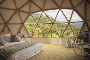 Un espacioso dormitorio con una cama grande, una mesita de noche y vistas a un paisaje exuberante a través de una estructura de ventana geométrica.