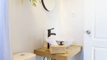 A compact bathroom with a toilet, a sink with a wooden shelf, a hanging plant, and a mirror on the wall.