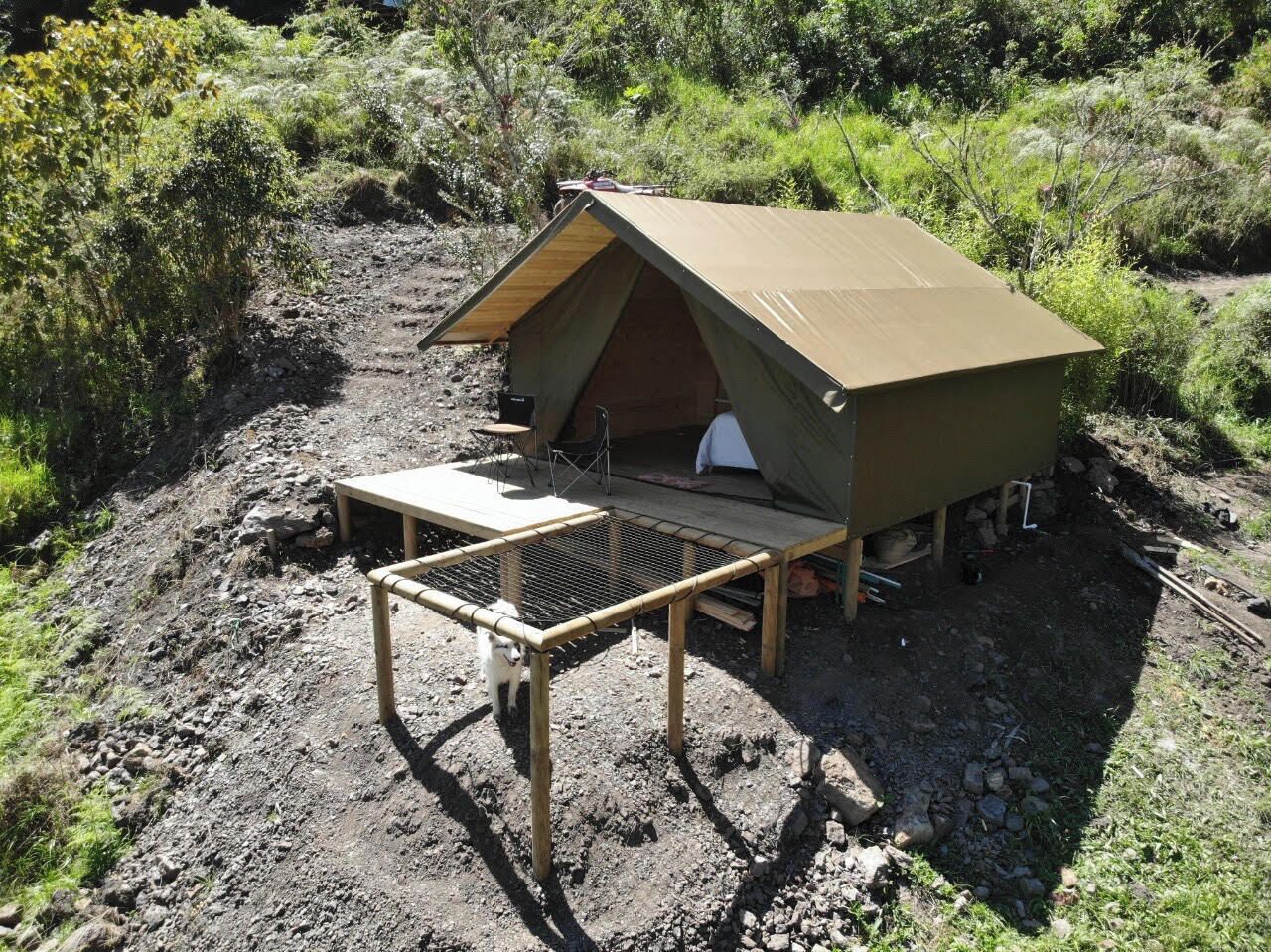 Una tenda petita d'una sola habitació amb una taula i cadires a l'exterior, envoltada per vegetació natural.
