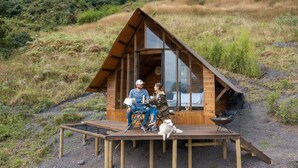 A wooden cabin with a sloped roof and large windows, situated on a dirt platform surrounded by grass and hills. - Cúspide Choachi Lodge (Choachí)