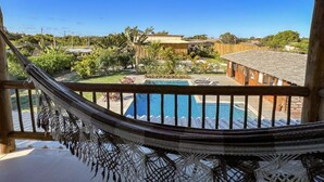 A hammock on a balcony overlooking a swimming pool with lounge chairs and a wooden building in the background. - Pousada Luz do Trevo (Porto Seguro)