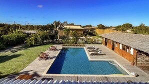 A rectangular pool with a wooden deck surrounding it, a thatched-roof building to the left, and a wooden deck with lounge chairs to the right. - Pousada Luz do Trevo (Porto Seguro)