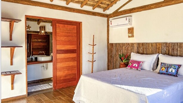 A bedroom with a wooden ceiling, a bed with a white bedspread, a bedside table, a nightstand, a wall-mounted air conditioner, and a wooden door leading to a bathroom. - Pousada Luz do Trevo (Porto Seguro)