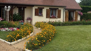 A two-story house with a tiled roof, wooden shutters, and a garden with flowers and a lawn.