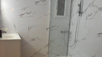 Une salle de bain moderne dotée d’une douche à parois de verre, de murs à motifs de marbre, d’un plancher en bois et d’un meuble-lavabo blanc.