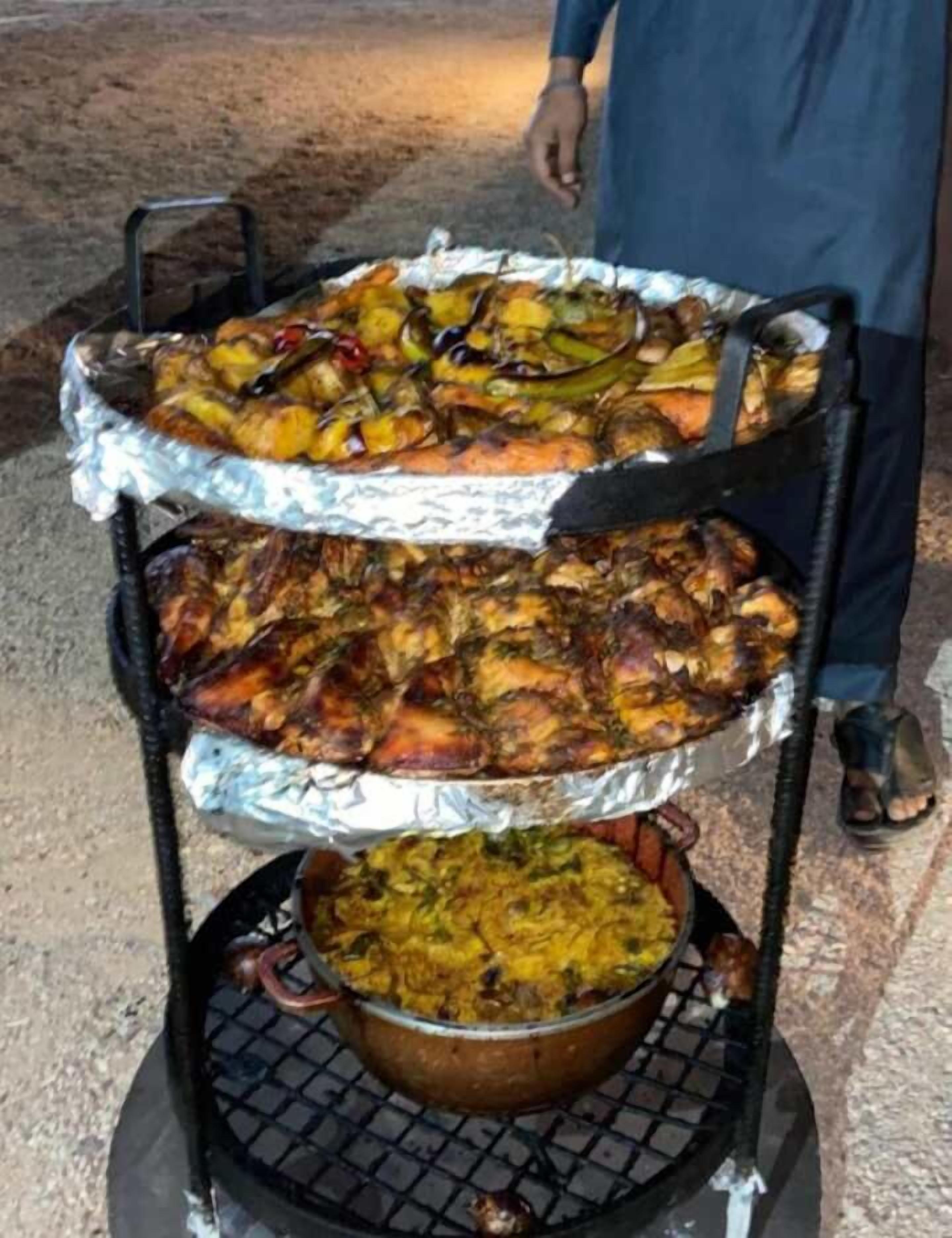 A large outdoor grill with multiple trays of food, including grilled meats and a pot of rice.