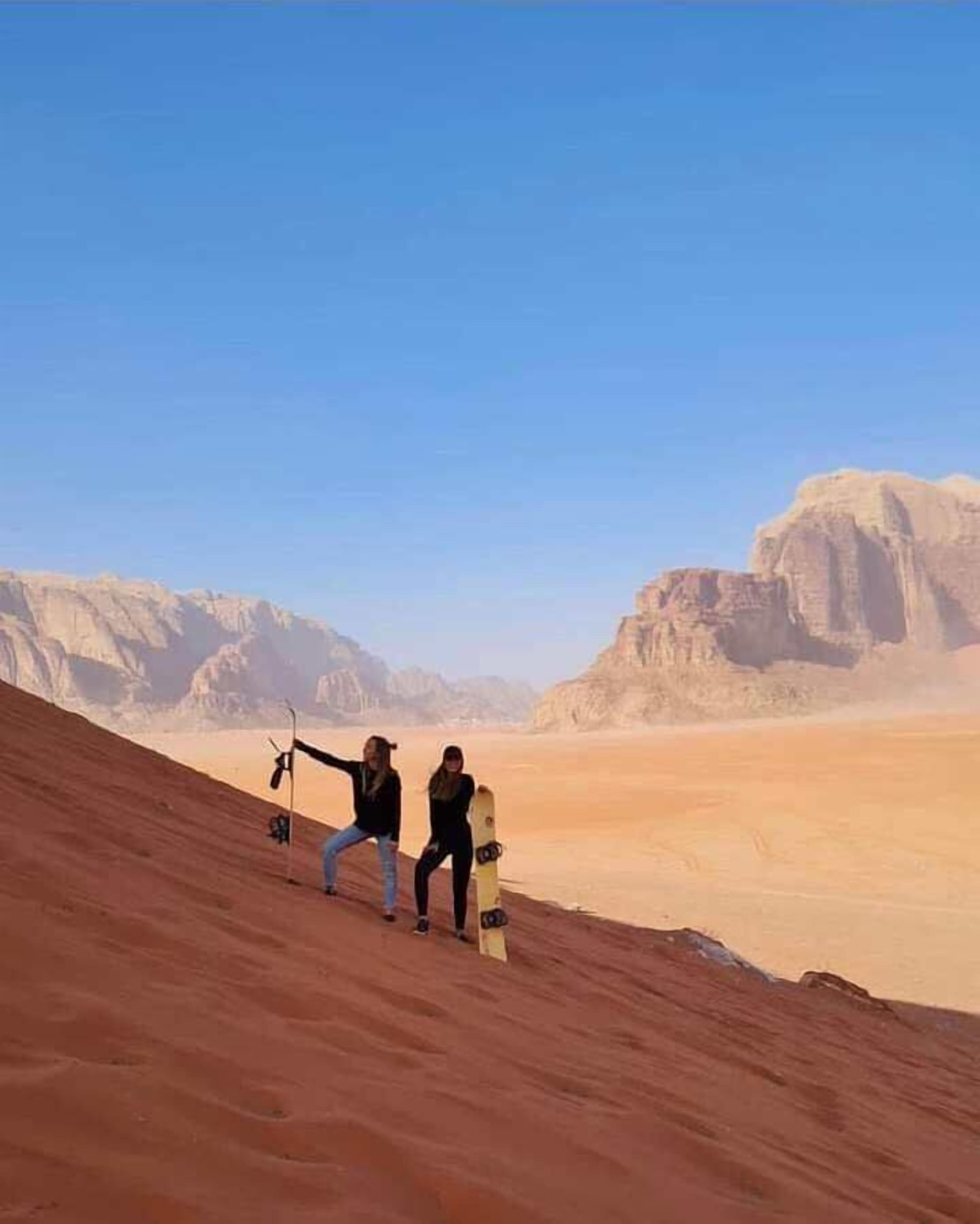 A desert landscape with two people standing on a sand dune, one holding a snowboard.