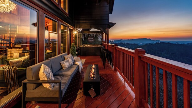 A wooden deck with a sofa, fire pit, and a view of a cityscape at sunset.