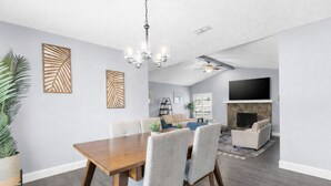 A modern living room with a fireplace, a dining area with a wooden table and chairs, and a large flat-screen TV mounted on the wall. - Leafy Lodge, Near Springs, Beach, Hot Tub, Fenced (Spring Hill)