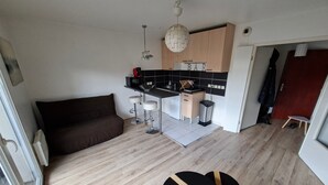 Un espacio habitable compacto con kitchenette, una zona de comedor con una mesa pequeña y taburetes, un sofá y una puerta que da a otra habitación.