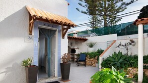 Una casa con patio empedrado, una puerta con un cartel, una silla y plantas en macetas.