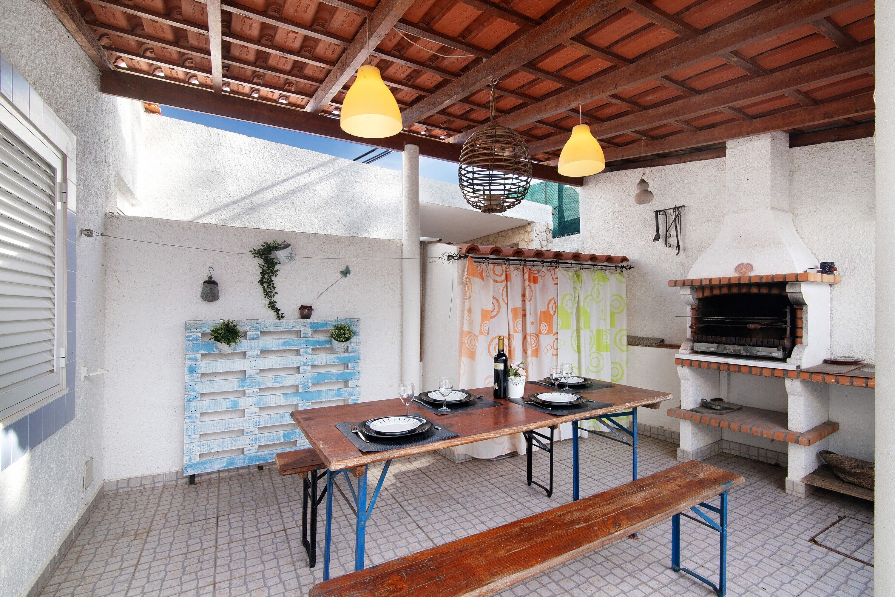An outdoor patio with a wooden table set for dining, a bench, a brick oven, and hanging lights.