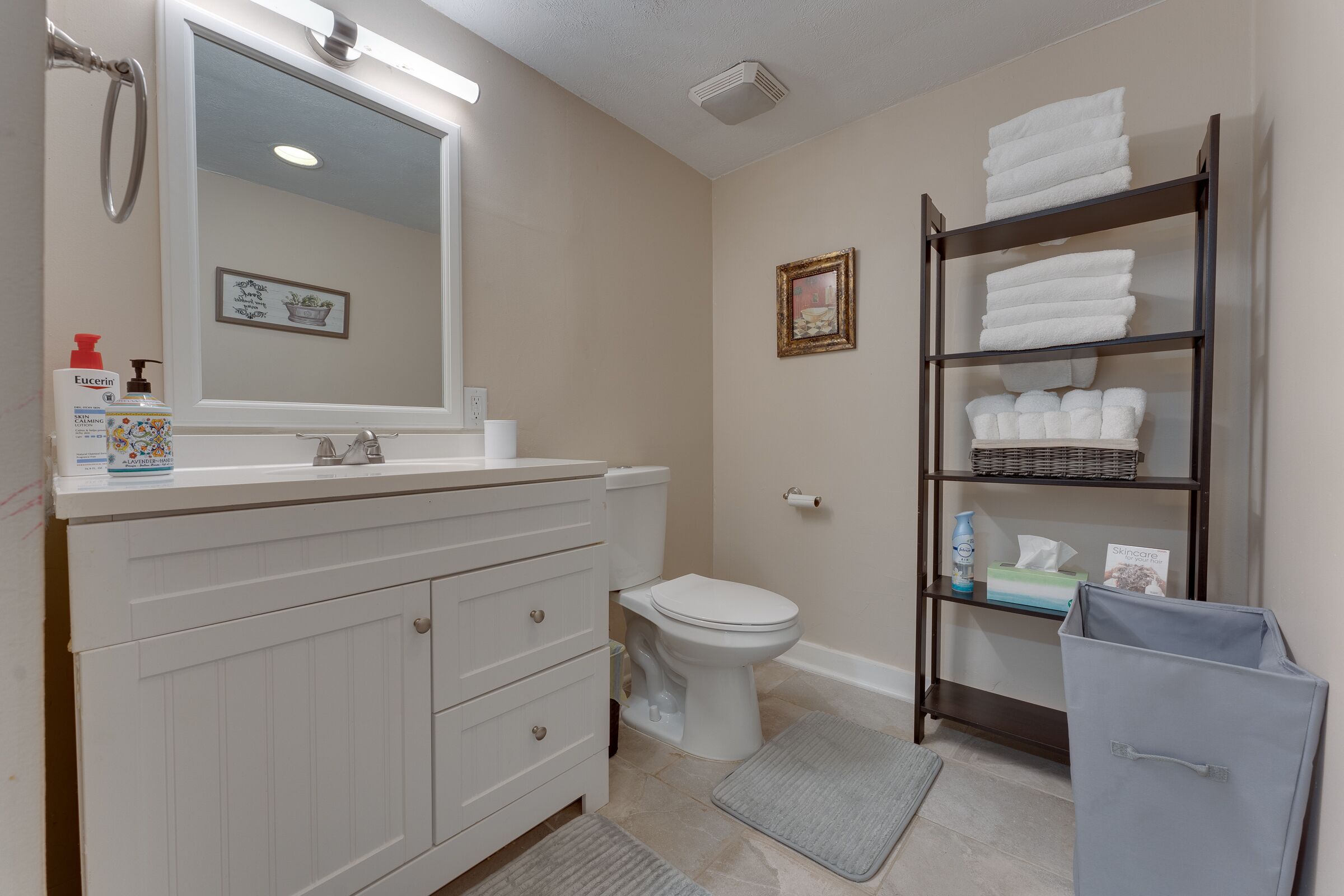 Une salle de bain avec un meuble-lavabo blanc, des toilettes, une étagère avec des serviettes et un tableau encadré au mur.