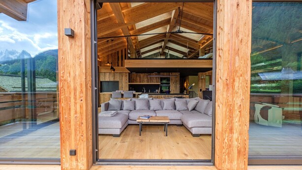 A modern wooden interior with a sectional sofa, a wooden coffee table, and a kitchen area visible through a glass door. - Chalet 'Aikoa' with Mountain View, Private Terrace and Wi-Fi (Chamonix-Mont-Blanc)