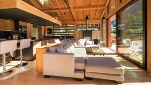 A modern interior with a wooden ceiling, a large sofa, a bar area, and large windows. - Chalet 'Aikoa' with Mountain View, Private Terrace and Wi-Fi (Chamonix-Mont-Blanc)