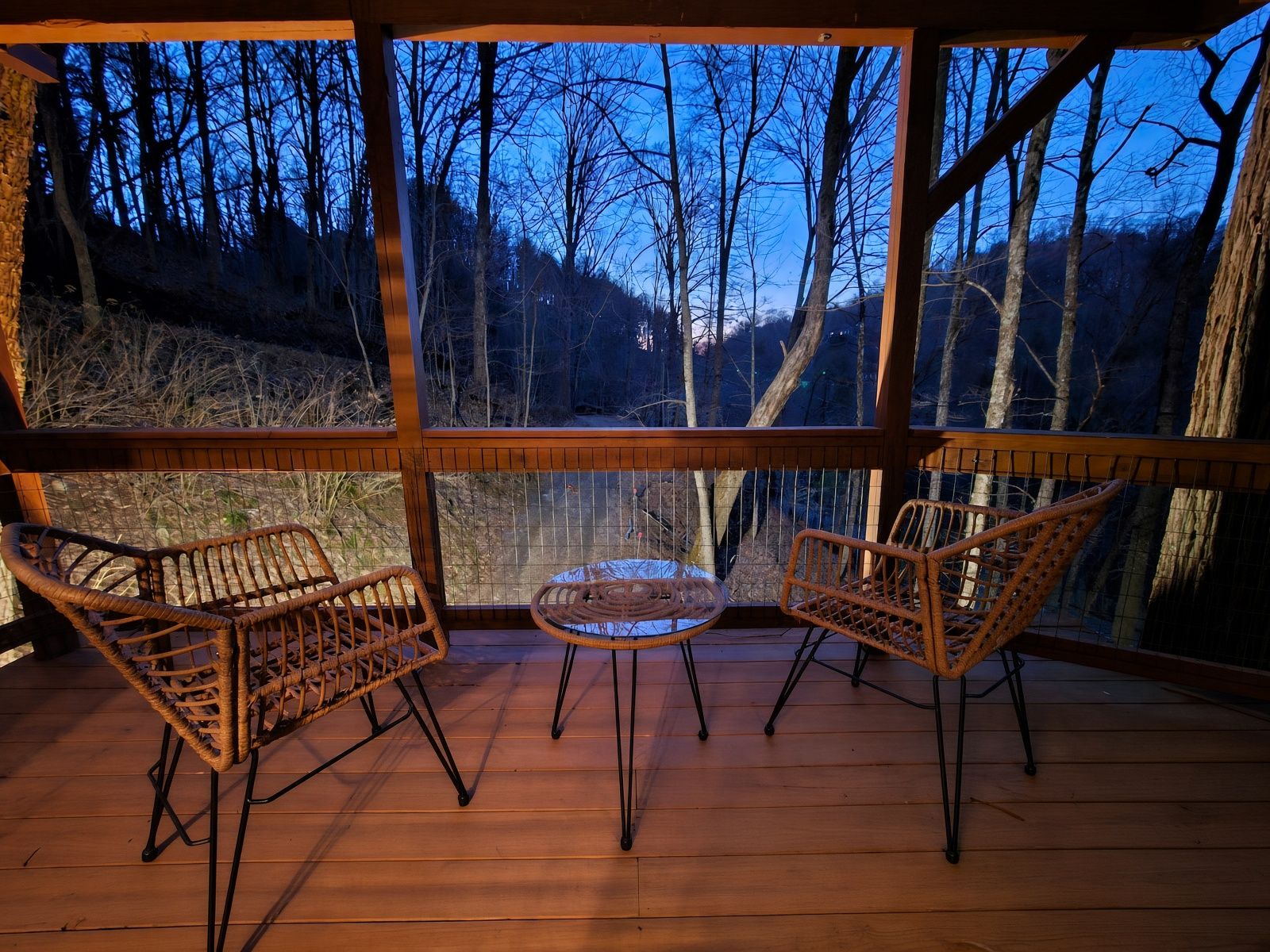 A wooden deck with wicker chairs and a small table, overlooking a forested area.