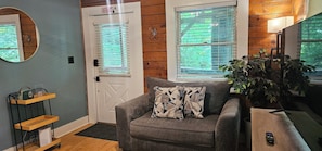 A room with a grey sofa, a wooden door with an exit sign, a mirror, a plant, and a TV on a wooden table. - Romantic Summer Couples Escape: Private Hot Tub + EV Charger + 600Mbps WiFi (Mars Hill)
