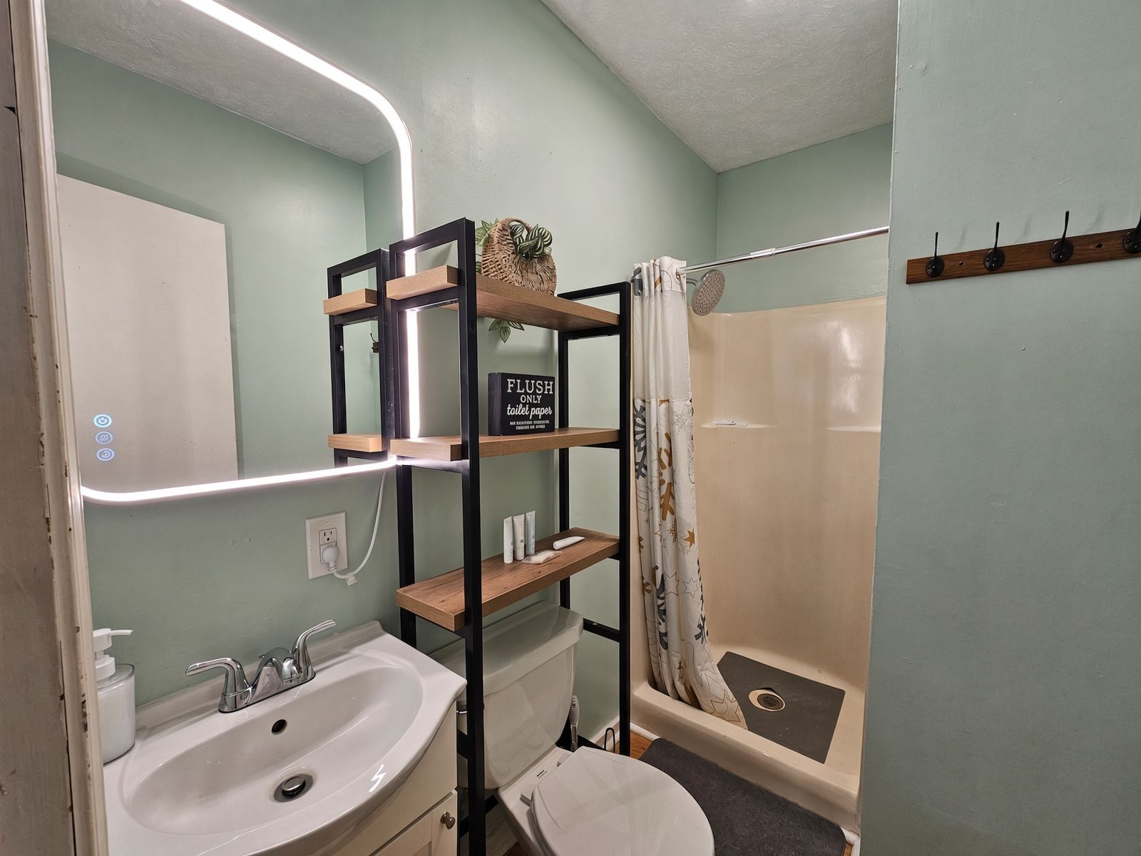 A bathroom with a sink, toilet, and shower. There is a shelf with toiletries and a sign that says 'FLUSH ONLY toilet paper'.