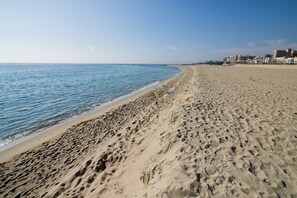 Una playa ancha de arena con huellas, un mar azul transparente y edificios al fondo.