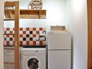 A compact kitchen with a washing machine, a refrigerator, and a wooden shelf above the sink. - Sycamore Cottage - UK45923 (Ballater)