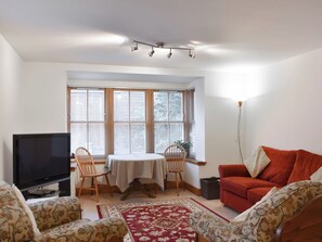 A living room with a flat-screen TV, two wooden chairs, a red sofa, a dining table with a white tablecloth, and a large window with blinds. - Sycamore Cottage - UK45923 (Ballater)