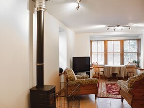 A living room with a black wood stove, a television, two patterned sofas, a dining table with chairs, and a large window with blinds. - Sycamore Cottage - UK45923 (Ballater)